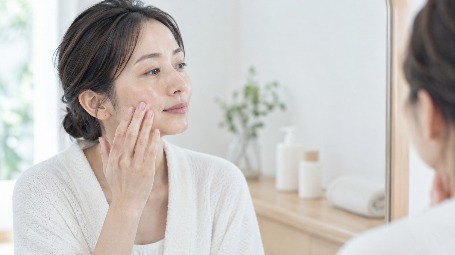 洗面台の鏡の前で、頬に手を添えて肌の状態を確かめている女性のイメージ写真。
